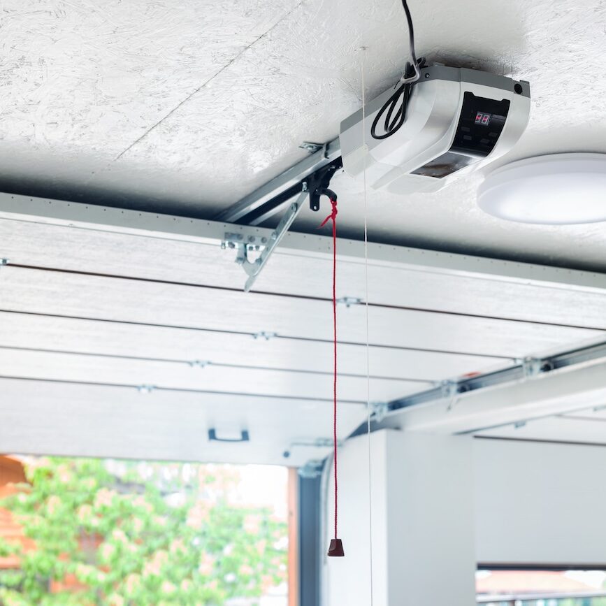Opening door and automatic garage door opener electric engine gear mounted on ceiling with emergency cord. Double place empty garage interior with rolling entrance gate.